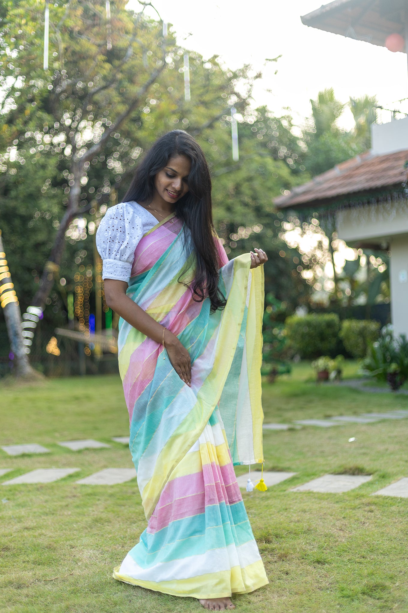 Woman in a colorful saree standing outdoors on grass with trees and a building in the background